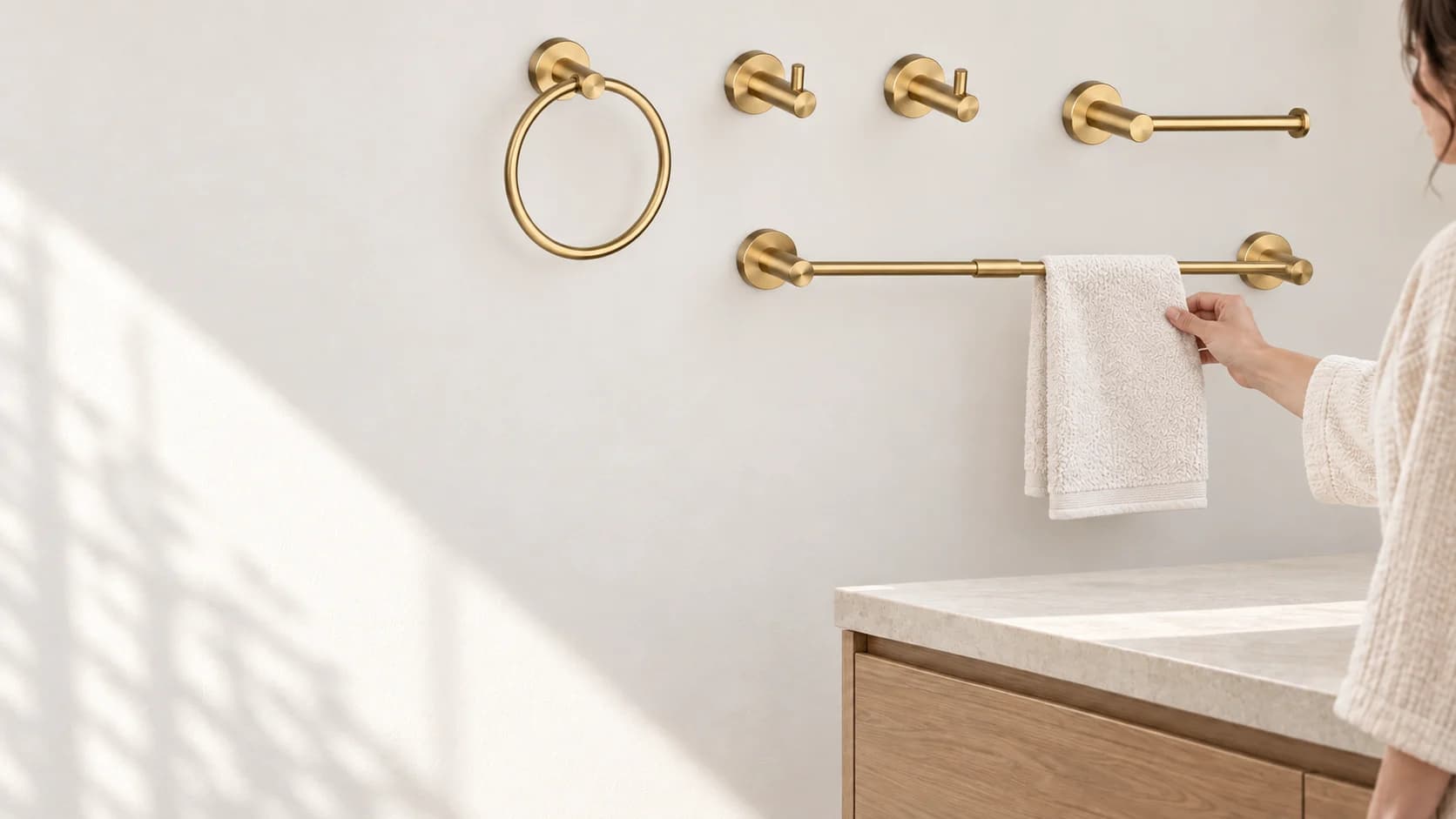 Una persona accanto al piano lavabo appende un asciugamano al set di ganci EYN BA1308, installato a parete, con finitura oro spazzolato.